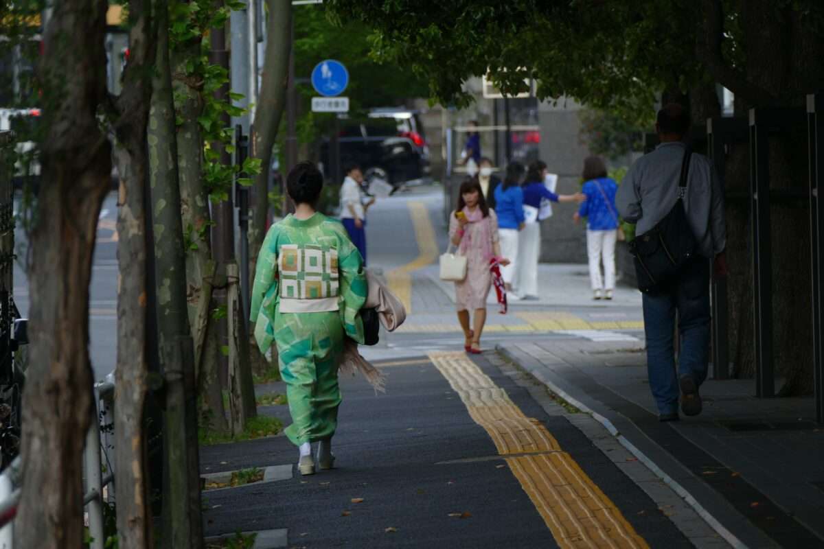 Personne vêtue d'un kimono traditionnel vert marchant sur un trottoir urbain avec des passants en arrière-plan.