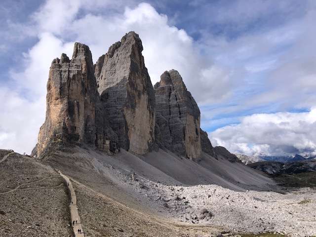 Vue impressionnante des Trois Cimes de Lavaredo avec leurs falaises escarpées se détachant sur un ciel dramatique, tandis que des randonneurs suivent le sentier à leur base dans les Dolomites, Italie.